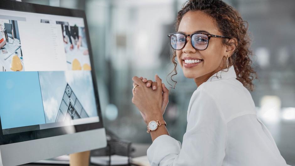 Eine lächelnde Frau mit Brille sitzt an einem Schreibtisch und schaut auf einen Computerbildschirm.