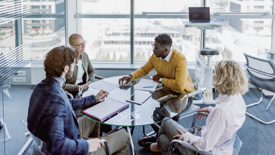 Vier Personen sitzen an einem runden Tisch in einem modernen Büro und diskutieren, während Notizblätter und ein Laptop auf dem Tisch liegen.