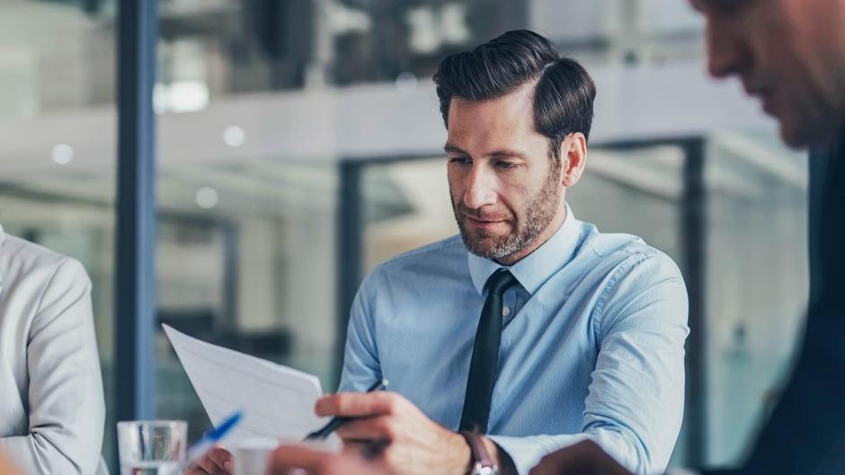 Mann in einem hellblauen Hemd und schwarzer Krawatte liest Dokumente in einem modernen Büro.