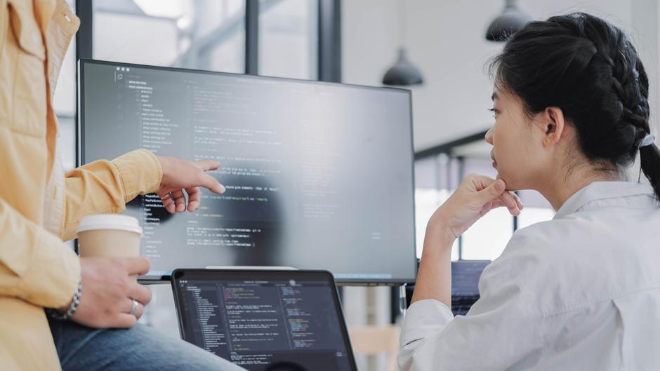 Zwei Personen diskutieren vor einem Computerbildschirm mit Code, eine Person zeigt auf den Bildschirm.