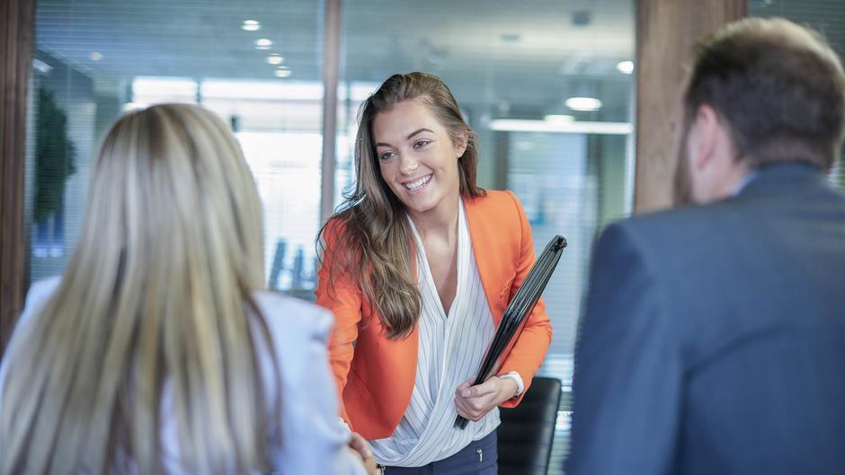 Eine lächelnde Frau in einem orangefarbenen Blazer schüttelt einer anderen Frau die Hand, während ein Mann im Hintergrund zuschaut.