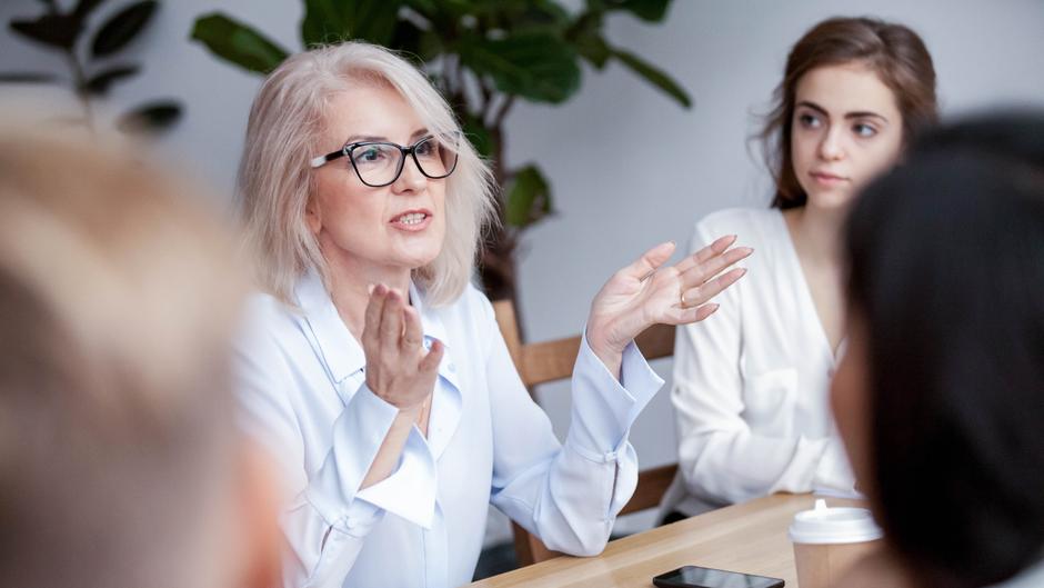 Eine blonde Frau mit Brille spricht leidenschaftlich in einer Besprechung, während eine junge Frau aufmerksam zuhört.